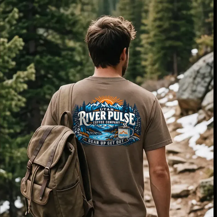 A hiker on a snow-edged mountain trail, seen from behind, wearing the Utah River Pulse Coffee Company tee with a canvas pack over one shoulder.